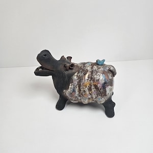 May include: A ceramic hippo figurine with a black head and legs, and a textured body with iridescent and colorful accents. A small blue bird sits on its back. The hippo has an open mouth and a smiling expression.