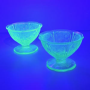 May include: Two vintage, light green glass dessert bowls with floral patterns. The bowls have a scalloped rim and sit on a pedestal base. The glass glows under a black light, creating a neon effect. The bowls are on a blue surface.