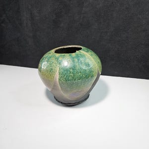 May include: A small, green and brown ceramic vase with a rounded shape and a wide opening. The vase has a speckled glaze and a slightly uneven surface.