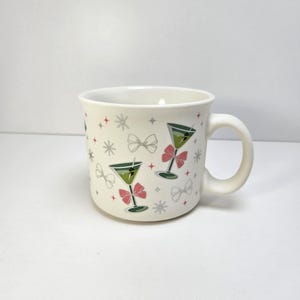May include: Off-white ceramic mug with a curved handle. The mug is decorated with martini glasses, pink bows, and silver starbursts. The martini glasses have green olives. The mug is on a white surface.