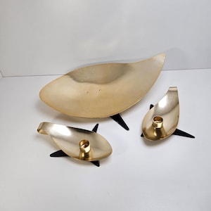 May include: A set of three gold-colored metal decorative pieces. The largest is a leaf-shaped bowl, and the other two are candle holders. All pieces have black, star-shaped bases. The items are displayed on a white surface.