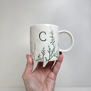 K&ouml;nnte beinhalten: Wei&szlig;er Keramikbecher mit schwarzem Buchstaben "C" und handgemaltem botanischen Design in Gr&uuml;n, Blau und Rosa. Der Becher hat einen gebogenen Henkel. Der Becher wird von einer Hand vor wei&szlig;em Hintergrund gehalten.