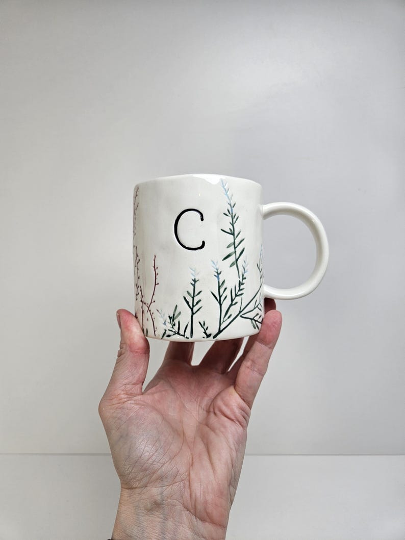 K&ouml;nnte beinhalten: Wei&szlig;er Keramikbecher mit schwarzem Buchstaben "C" und handgemaltem botanischen Design in Gr&uuml;n, Blau und Rosa. Der Becher hat einen gebogenen Henkel. Der Becher wird von einer Hand vor wei&szlig;em Hintergrund gehalten.