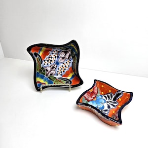 May include: Two decorative, hand-painted ceramic dishes. The larger dish has a black rim and a colorful floral design with orange, blue, and white accents. The smaller dish has a similar design with an orange background.