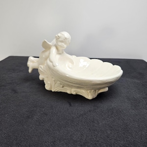 May include: A cream-colored ceramic dish shaped like a shell, featuring a sculpted cherub figure with wings attached to the side. The base of the dish has ornate, carved detailing, and the entire piece has a smooth, glossy finish throughout.