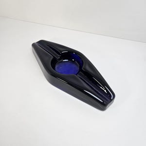 May include: A black ashtray with a deep blue interior. The ashtray has a sleek, geometric design with a central circular depression and two elongated grooves for resting cigarettes. The glossy finish reflects light, highlighting its shape.