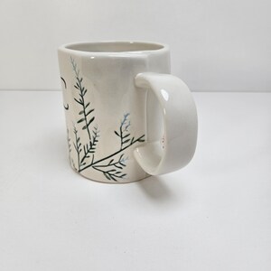 K&ouml;nnte beinhalten: Wei&szlig;er Keramikbecher mit gebogenem Henkel. Der Becher hat ein handgemaltes Design aus gr&uuml;nen und blauen Blumenzweigen. Der Becher hat eine glatte, gl&auml;nzende Oberfl&auml;che und ist vor einem wei&szlig;en Hintergrund platziert. Der Becher hat eine Standardgr&ouml;&szlig;e f&uuml;r Kaffee oder Tee.
