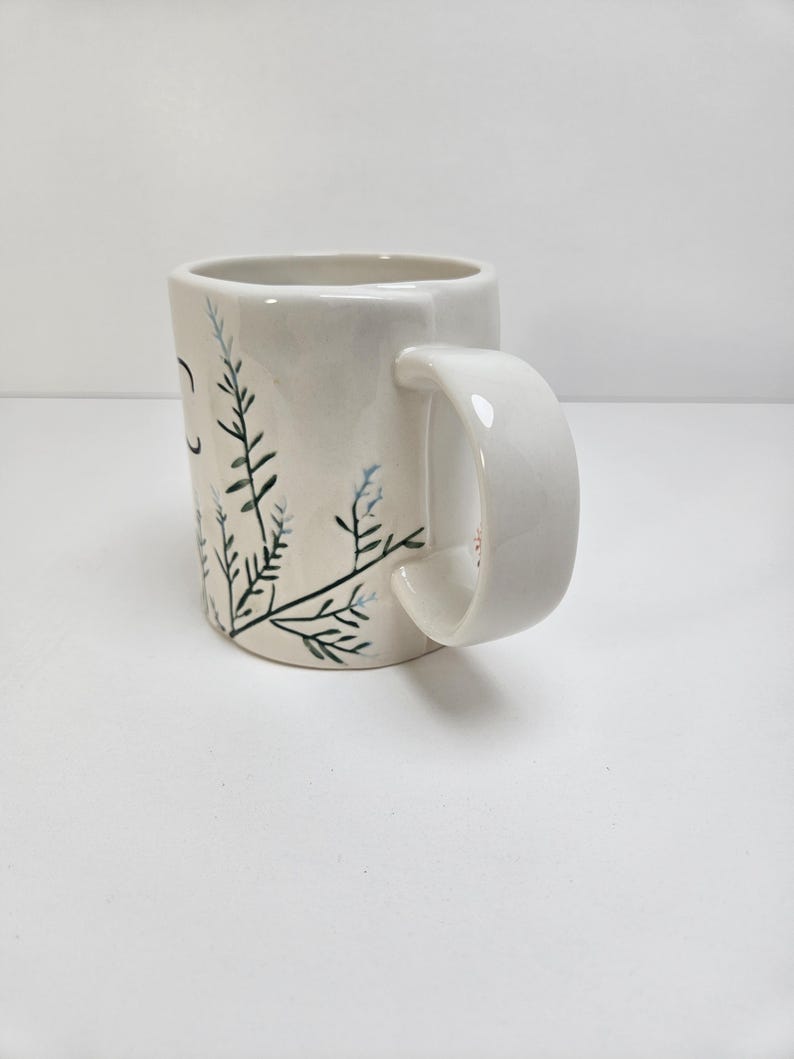 K&ouml;nnte beinhalten: Wei&szlig;er Keramikbecher mit gebogenem Henkel. Der Becher hat ein handgemaltes Design aus gr&uuml;nen und blauen Blumenzweigen. Der Becher hat eine glatte, gl&auml;nzende Oberfl&auml;che und ist vor einem wei&szlig;en Hintergrund platziert. Der Becher hat eine Standardgr&ouml;&szlig;e f&uuml;r Kaffee oder Tee.