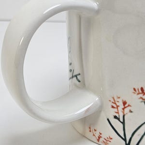 K&ouml;nnte beinhalten: Cremefarbene Keramik-Tasse mit gebogenem Henkel. Die Tasse hat ein Blumendesign mit roten und schwarzen Akzenten. Der Griff ist glatt und gl&auml;nzend, und die Tasse scheint handgefertigt zu sein.