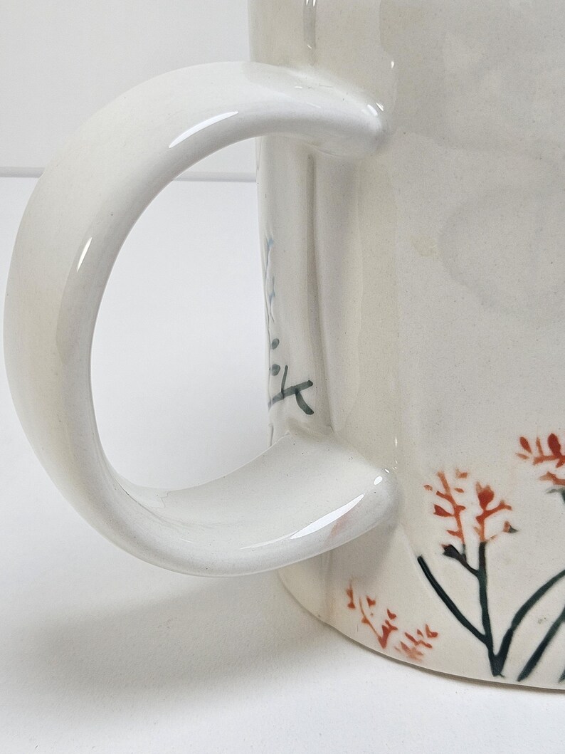 K&ouml;nnte beinhalten: Cremefarbene Keramik-Tasse mit gebogenem Henkel. Die Tasse hat ein Blumendesign mit roten und schwarzen Akzenten. Der Griff ist glatt und gl&auml;nzend, und die Tasse scheint handgefertigt zu sein.