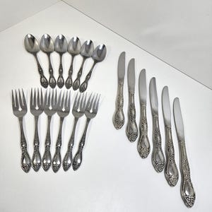 May include: A collection of vintage silverware, including spoons, forks, and knives. The silverware has ornate floral detailing on the handles and a shiny, metallic finish. The arrangement is set against a white background.