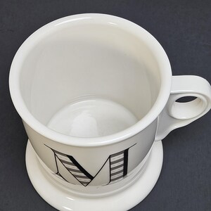 May include: White ceramic mug with a large letter "M" in black and gray stripes. The mug has a rounded base and a handle with a circular opening. The mug sits on a matching white saucer.