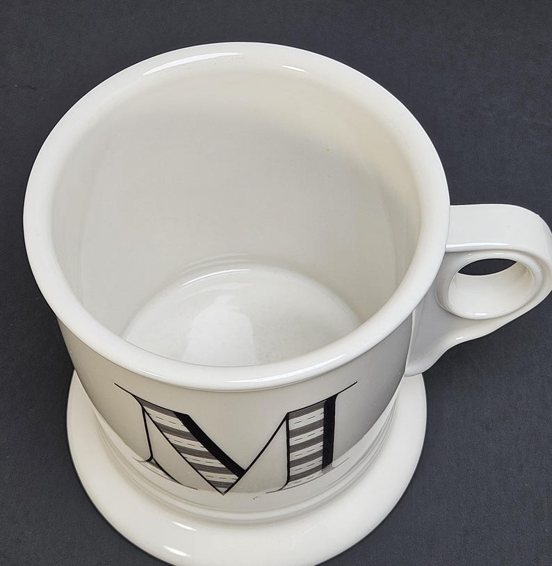 May include: White ceramic mug with a large letter "M" in black and gray stripes. The mug has a rounded base and a handle with a circular opening. The mug sits on a matching white saucer.