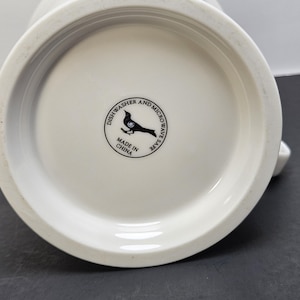 May include: White ceramic mug with a rounded base. The bottom of the mug features a circular stamp with a bird silhouette and the text "Dishwasher and Microwave Safe". The mug is made in China.