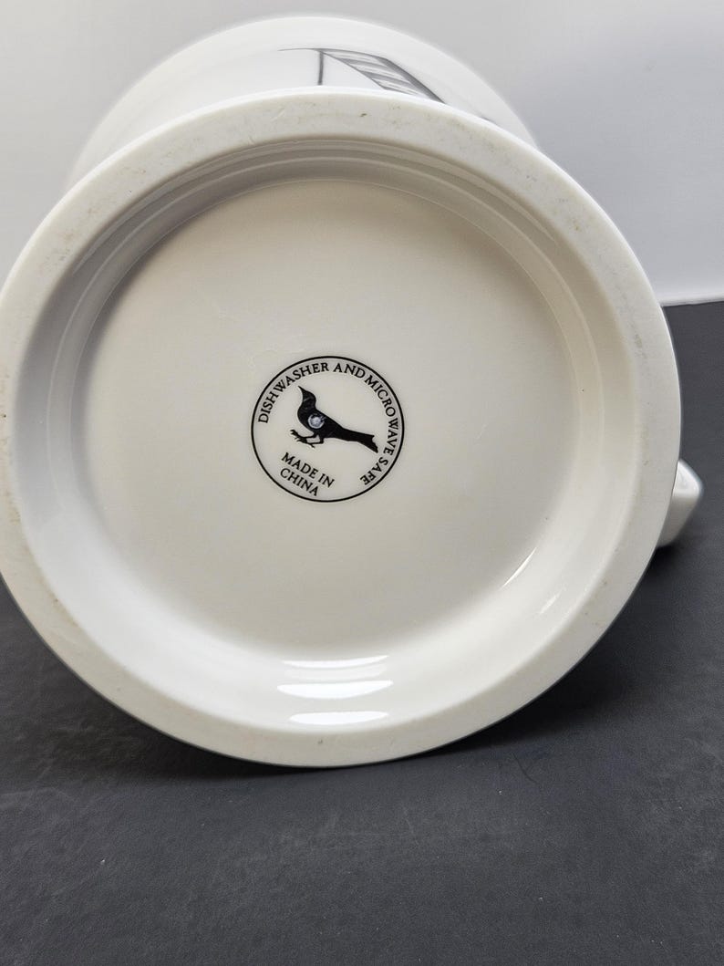 May include: White ceramic mug with a rounded base. The bottom of the mug features a circular stamp with a bird silhouette and the text "Dishwasher and Microwave Safe". The mug is made in China.