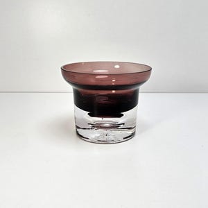 May include: A clear glass votive candle holder with a wide, dark purple rim and a dark purple base. The bottom half of the holder is clear, allowing light to pass through. The candle holder is designed with a modern aesthetic.