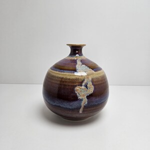 May include: A handcrafted ceramic vase with a rounded body and a narrow neck. The vase features a dark brown base with bands of blue and tan, and a light-colored, abstract design. The vase is approximately 6 inches tall.