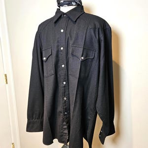 May include: A black long-sleeve shirt with two chest pockets and silver snap closures. A black and white bandana is draped around the collar. The shirt is displayed on a mannequin.