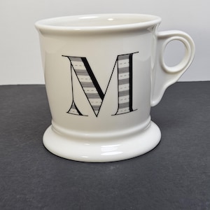 May include: White ceramic mug with a black letter "M" on the front. The letter is stylized with horizontal lines. The mug has a handle and a wide, rounded base. The mug is on a dark gray surface.