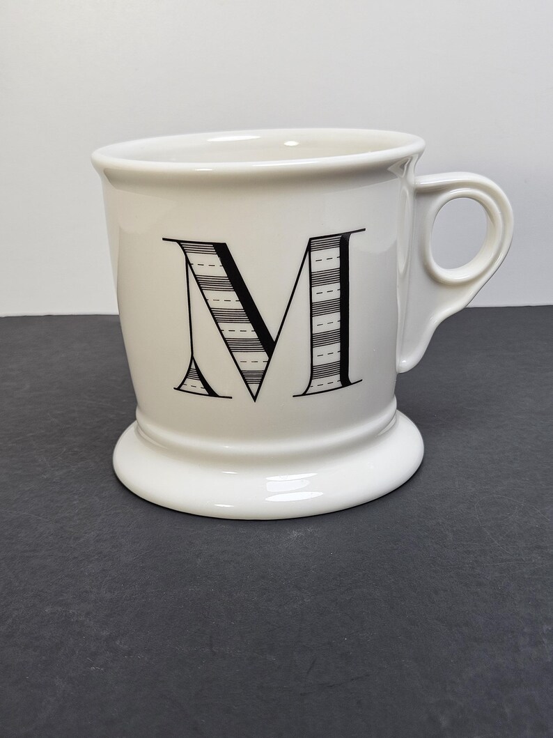 May include: White ceramic mug with a black letter "M" on the front. The letter is stylized with horizontal lines. The mug has a handle and a wide, rounded base. The mug is on a dark gray surface.