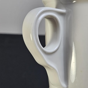 May include: Close-up of a white ceramic mug handle. The handle is a curved, loop design, attached to the side of the mug. The mug has a glossy finish and a smooth surface. The background is a dark gray.