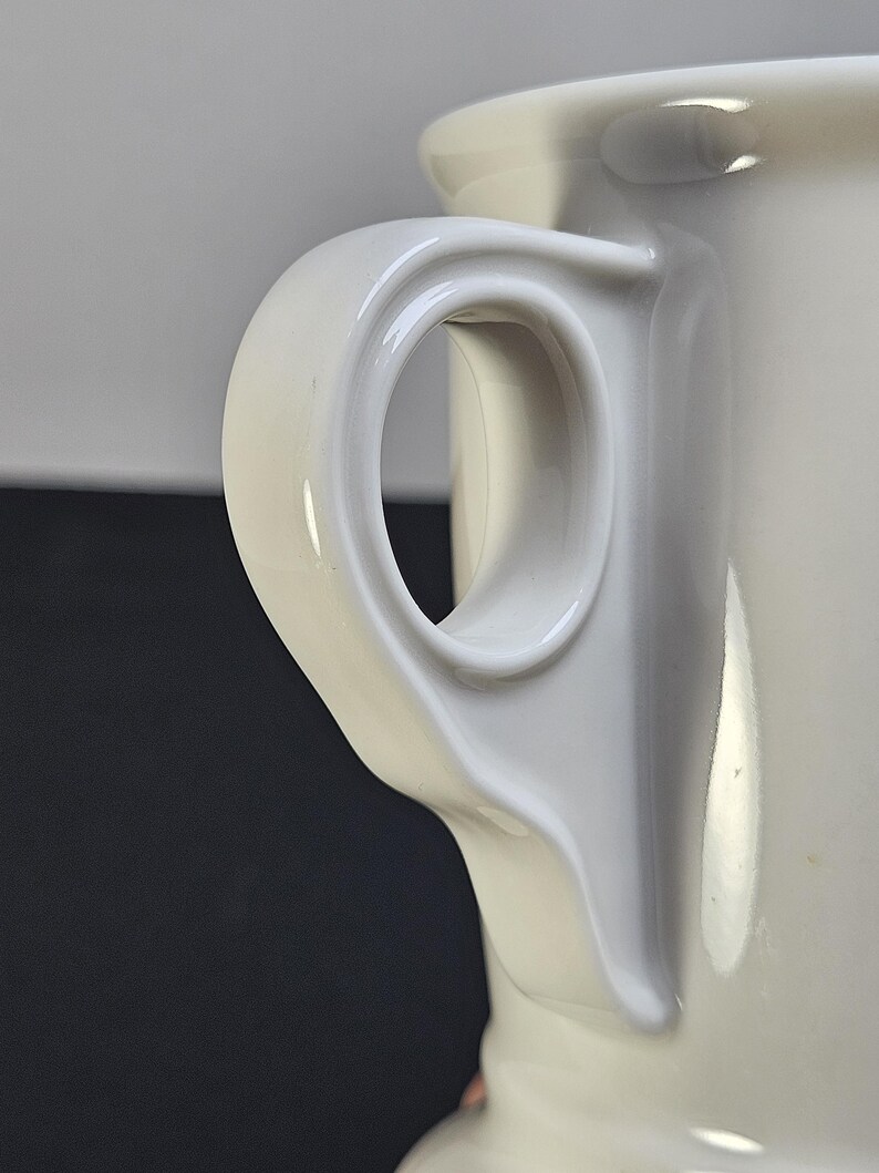 May include: Close-up of a white ceramic mug handle. The handle is a curved, loop design, attached to the side of the mug. The mug has a glossy finish and a smooth surface. The background is a dark gray.