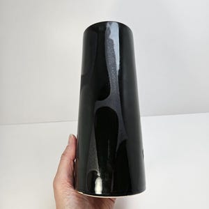 May include: A tall, black ceramic vase with a glossy finish. The vase has a slightly flared shape and a smooth, reflective surface. The black glaze has subtle variations in tone, adding depth to the design. The vase is held by a hand against a white background.