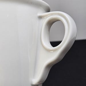 May include: Close-up of a white ceramic mug with a distinctive handle. The handle has a rounded, oval opening and is smoothly integrated into the mug's body. The mug's surface is glossy and reflects light.