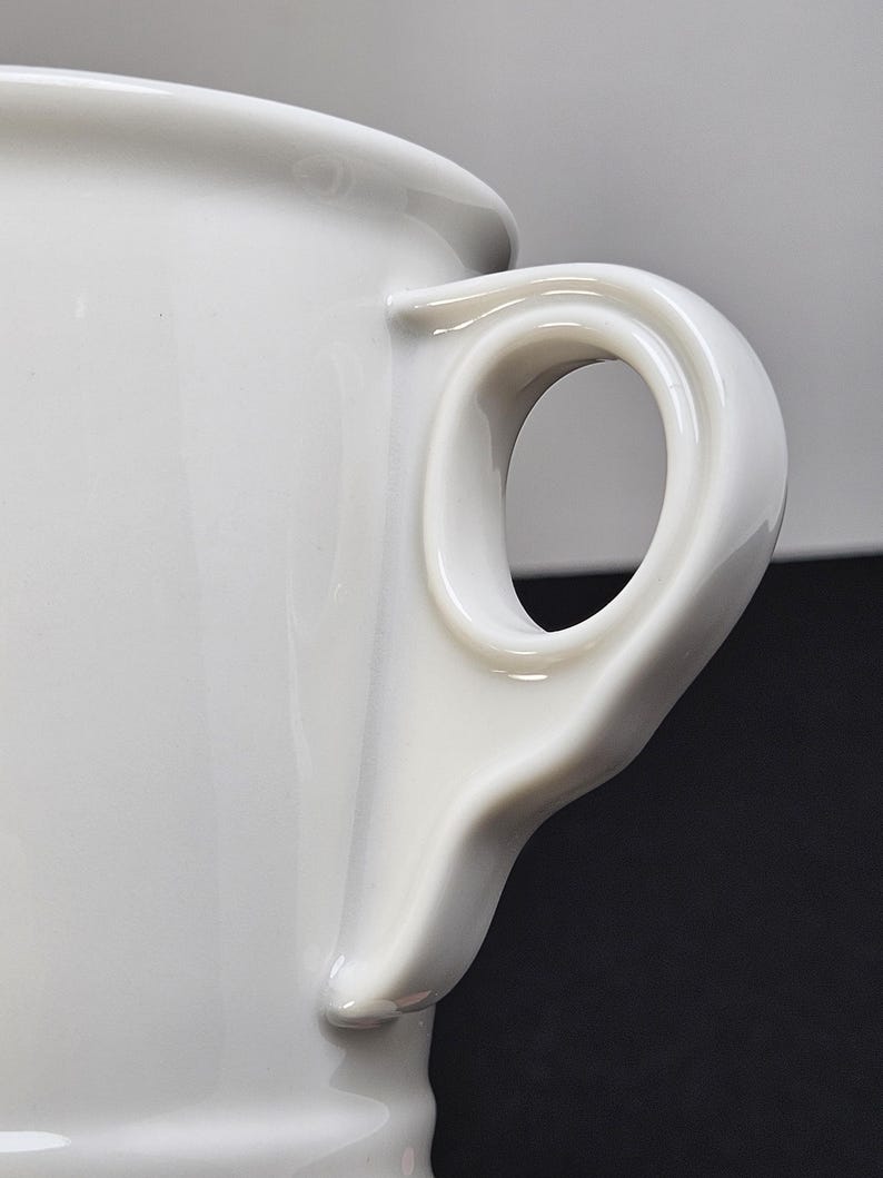 May include: Close-up of a white ceramic mug with a distinctive handle. The handle has a rounded, oval opening and is smoothly integrated into the mug's body. The mug's surface is glossy and reflects light.