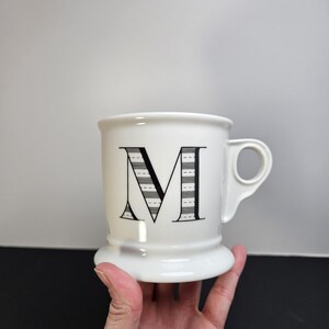 May include: White ceramic mug with a large black letter "M" on the front. The letter is decorated with a striped design. The mug has a handle and a wide base. The mug is held by a hand.