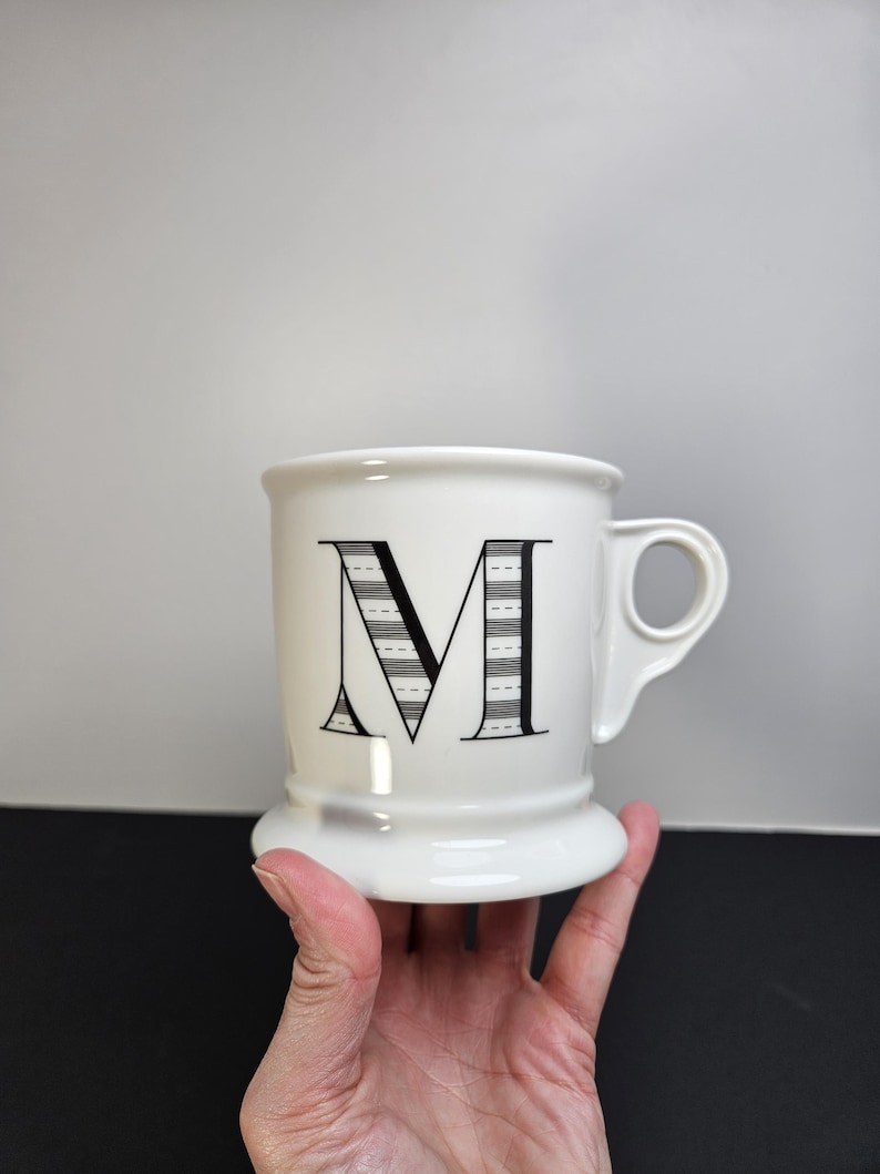 May include: White ceramic mug with a large black letter "M" on the front. The letter is decorated with a striped design. The mug has a handle and a wide base. The mug is held by a hand.