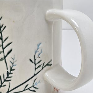 K&ouml;nnte beinhalten: Cremefarbene Keramik-Tasse mit gebogenem Henkel. Die Tasse hat ein handgemaltes botanisches Design mit dunkelgr&uuml;nen und hellblauen Bl&auml;ttern und Stielen. Die Tasse hat eine quadratische Form und eine glatte, gl&auml;nzende Oberfl&auml;che.