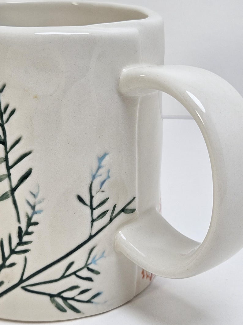 K&ouml;nnte beinhalten: Cremefarbene Keramik-Tasse mit gebogenem Henkel. Die Tasse hat ein handgemaltes botanisches Design mit dunkelgr&uuml;nen und hellblauen Bl&auml;ttern und Stielen. Die Tasse hat eine quadratische Form und eine glatte, gl&auml;nzende Oberfl&auml;che.