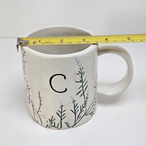 K&ouml;nnte beinhalten: Wei&szlig;er Keramikbecher mit schwarzem Buchstaben "C" und bemaltem botanischen Design. Der Becher ist etwa 10 cm breit, gemessen mit einem gelben Ma&szlig;band. Der Becher hat einen Henkel und ist mit gr&uuml;nen, blauen und roten Pflanzenillustrationen verziert.