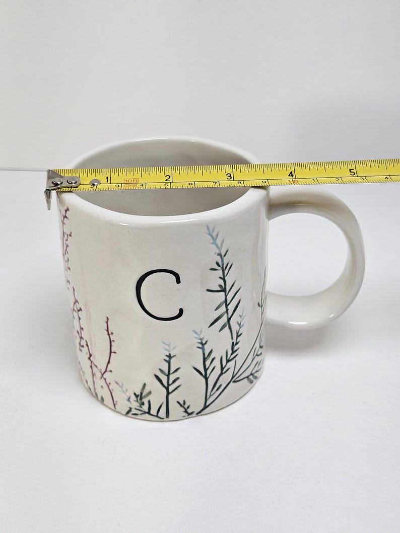 K&ouml;nnte beinhalten: Wei&szlig;er Keramikbecher mit schwarzem Buchstaben "C" und bemaltem botanischen Design. Der Becher ist etwa 10 cm breit, gemessen mit einem gelben Ma&szlig;band. Der Becher hat einen Henkel und ist mit gr&uuml;nen, blauen und roten Pflanzenillustrationen verziert.