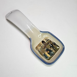 May include: A white ceramic spoon rest with a blue rim. The spoon rest features a colorful illustration of a group of people standing in front of a house.