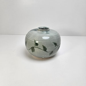 May include: A round, light blue ceramic vase with a small opening at the top. The vase features a delicate green vine and leaf design. The vase is a decorative item.