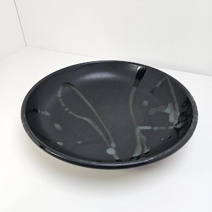 May include: A black ceramic serving bowl with a wide, shallow design. The bowl features a glossy finish with abstract, light gray splatter patterns. The rim is slightly raised, and the bowl is set against a white background.