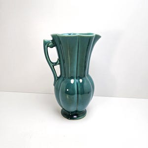 May include: A teal-colored ceramic pitcher with a handle and a flared, scalloped rim. The pitcher has a bulbous body and a pedestal base. The surface is glossy and reflects light, highlighting the vertical ribbed design. A decorative piece for the home.