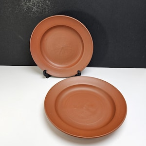 May include: Two round terracotta plates with a smooth, matte finish and a wide, flat rim. One plate is positioned upright on a small black stand, while the second plate lies flat in the foreground, showcasing the consistent reddish-brown color.