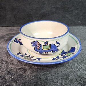 May include: A white ceramic bowl with a blue rim and a blue, green, and yellow design of animals. The bowl is sitting on a matching saucer.