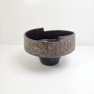 May include: A ceramic bowl with a dark brown, textured exterior featuring vertical grooves and white speckles. The rim has an asymmetrical, stepped design with a cutout section. The base is a smooth, solid black, tapered pedestal shape.