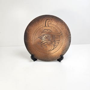 May include: A circular copper plate featuring a dark, hammered finish and a central engraved design of an eagle head in a stylized form. The plate is displayed on a small black plastic stand, showcasing a gradient of brown and bronze tones.