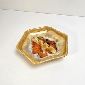 May include: A hexagonal ceramic ashtray with a tan exterior and a vibrant orange and brown interior design. The ashtray features three cigarette rests and a glossy finish, perfect for a retro or vintage aesthetic.