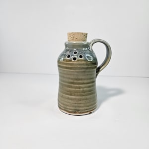 May include: A small, hand-thrown ceramic jar with a cork lid and a handle. The jar has a light green glaze with brown stripes and a few holes in the side.