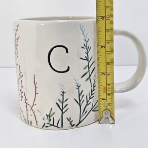 K&ouml;nnte beinhalten: Cremefarbene Keramik-Tasse mit schwarzem Buchstaben "C" und bemalten botanischen Akzenten in Gr&uuml;n, Blau und Burgunderrot. Die Tasse ist etwa 10 cm hoch, mit einem wei&szlig;en Henkel.