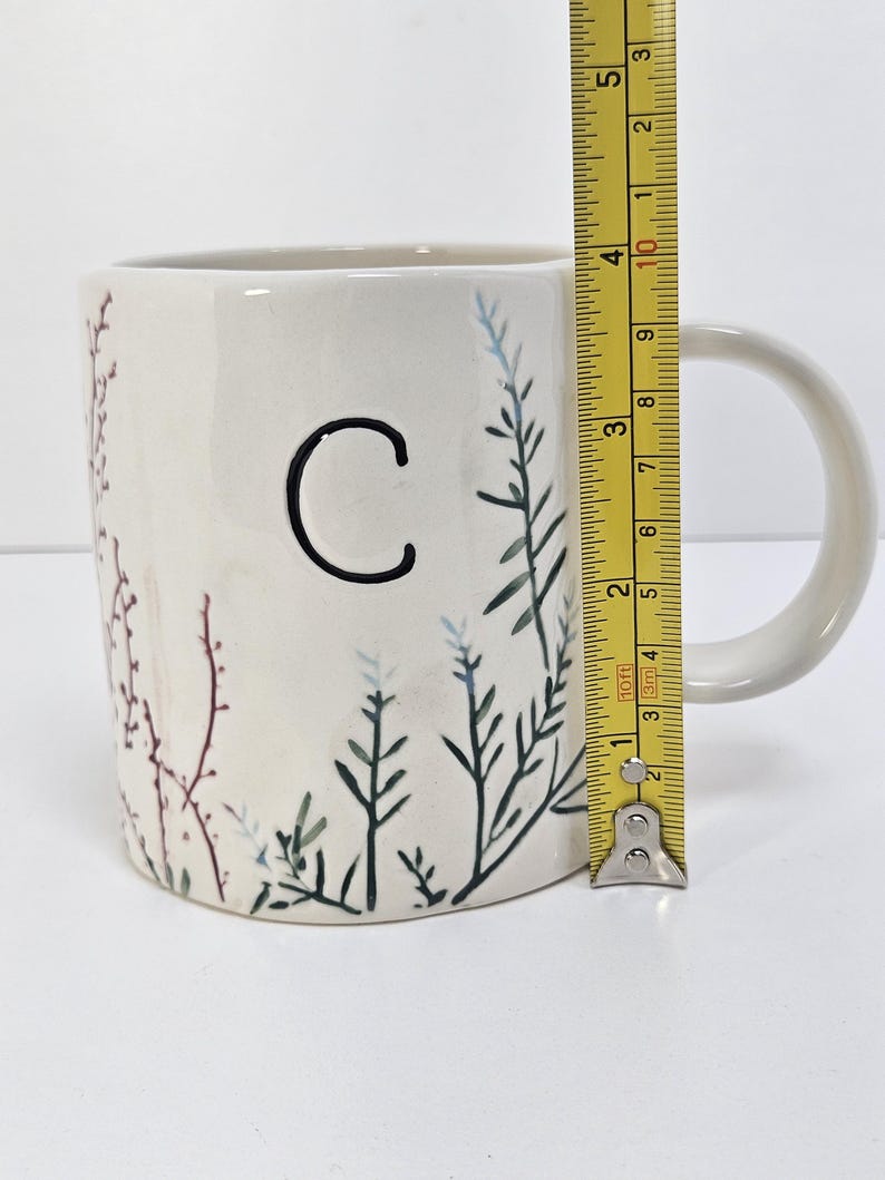 K&ouml;nnte beinhalten: Cremefarbene Keramik-Tasse mit schwarzem Buchstaben "C" und bemalten botanischen Akzenten in Gr&uuml;n, Blau und Burgunderrot. Die Tasse ist etwa 10 cm hoch, mit einem wei&szlig;en Henkel.