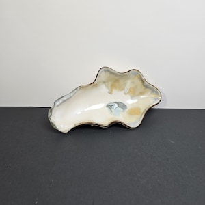 May include: An irregular, organic-shaped ceramic bowl with a glossy finish. The interior features a blend of cream, soft tan, and muted blue-grey glazes. The rim is defined by a dark, contrasting edge that follows the undulating, wavy contour.