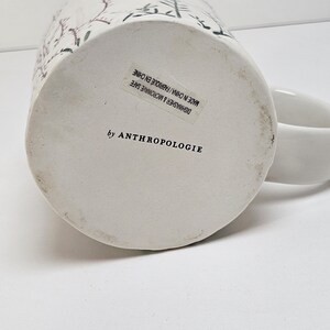 K&ouml;nnte beinhalten: Wei&szlig;er Keramikbecher mit floralem Design und Henkel. Der Boden des Bechers hat einen Aufkleber mit der Aufschrift "by ANTHROPOLOGIE" und "DISHWASHER & MICROWAVE SAFE MADE IN CHINA / FABRIQUE EN CHINE".