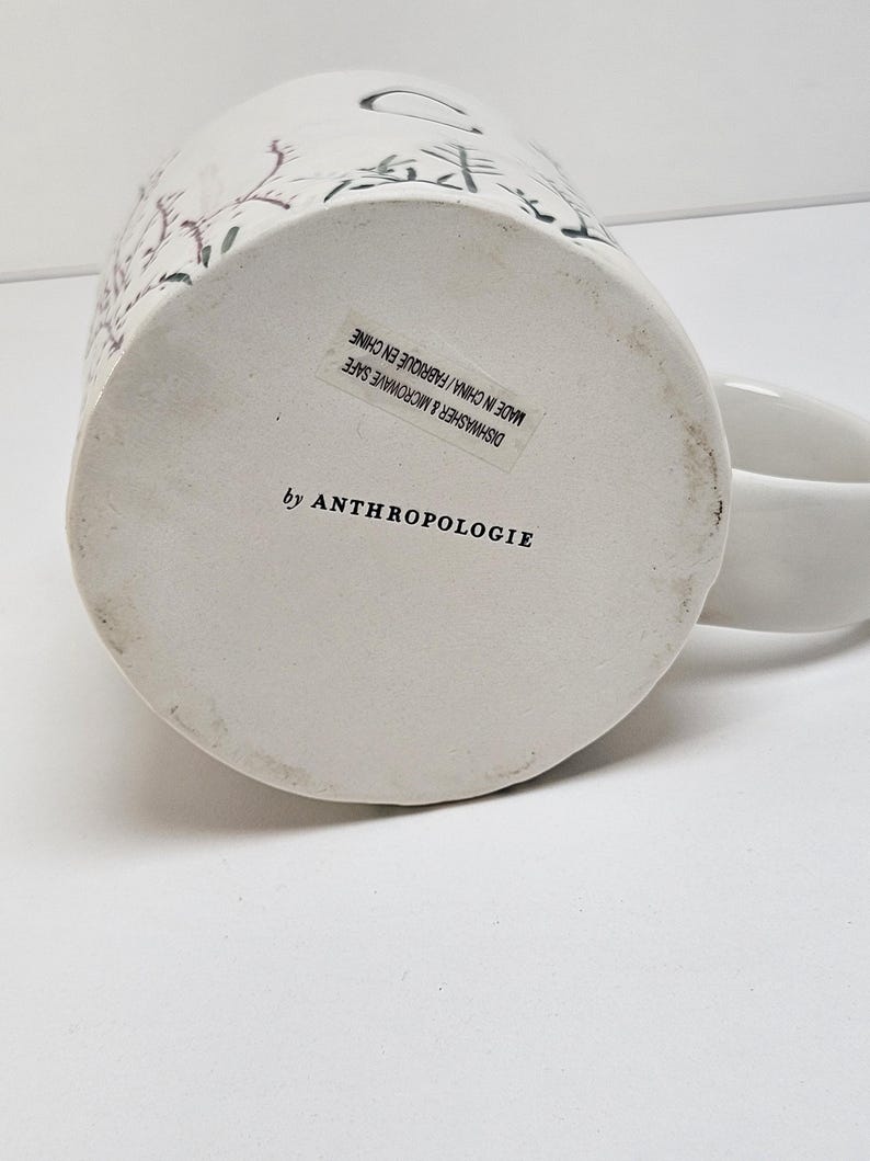 K&ouml;nnte beinhalten: Wei&szlig;er Keramikbecher mit floralem Design und Henkel. Der Boden des Bechers hat einen Aufkleber mit der Aufschrift "by ANTHROPOLOGIE" und "DISHWASHER & MICROWAVE SAFE MADE IN CHINA / FABRIQUE EN CHINE".